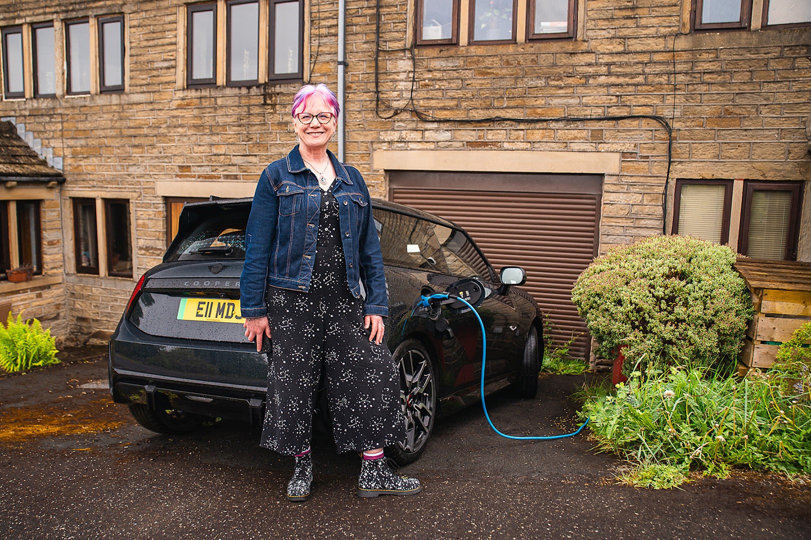 Woman standing beside a black MINI Electric plugged into a home charger outside a house